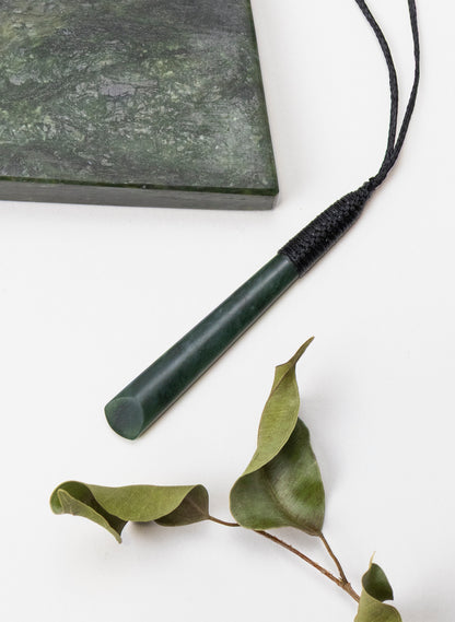 Long Kawakawa pounamu toki pendant beside a stone slab and leaves