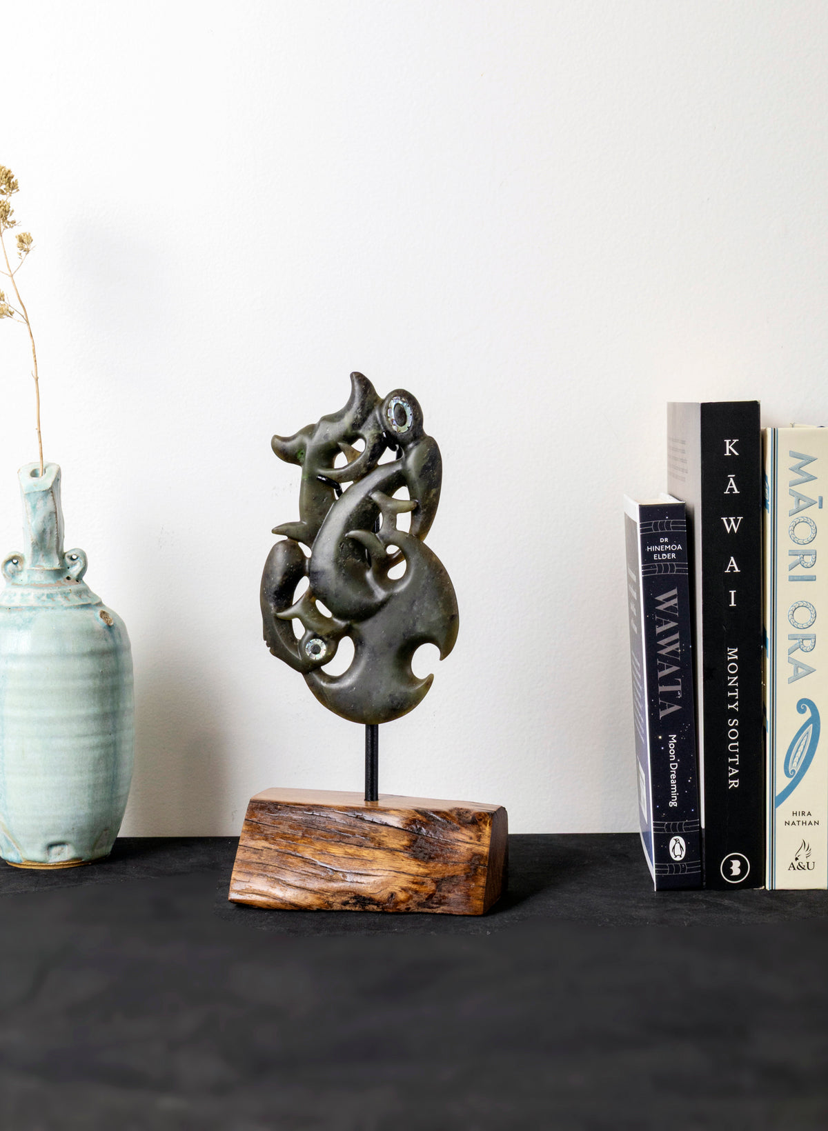 Manaia Pounamu on a Stand, displayed on a dark surface beside a pale vase and books
