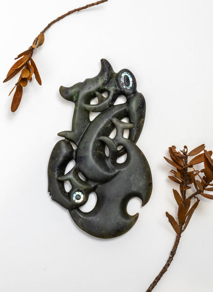 Manaia Pounamu on a Stand, carving laid flat beside dried leafy branches